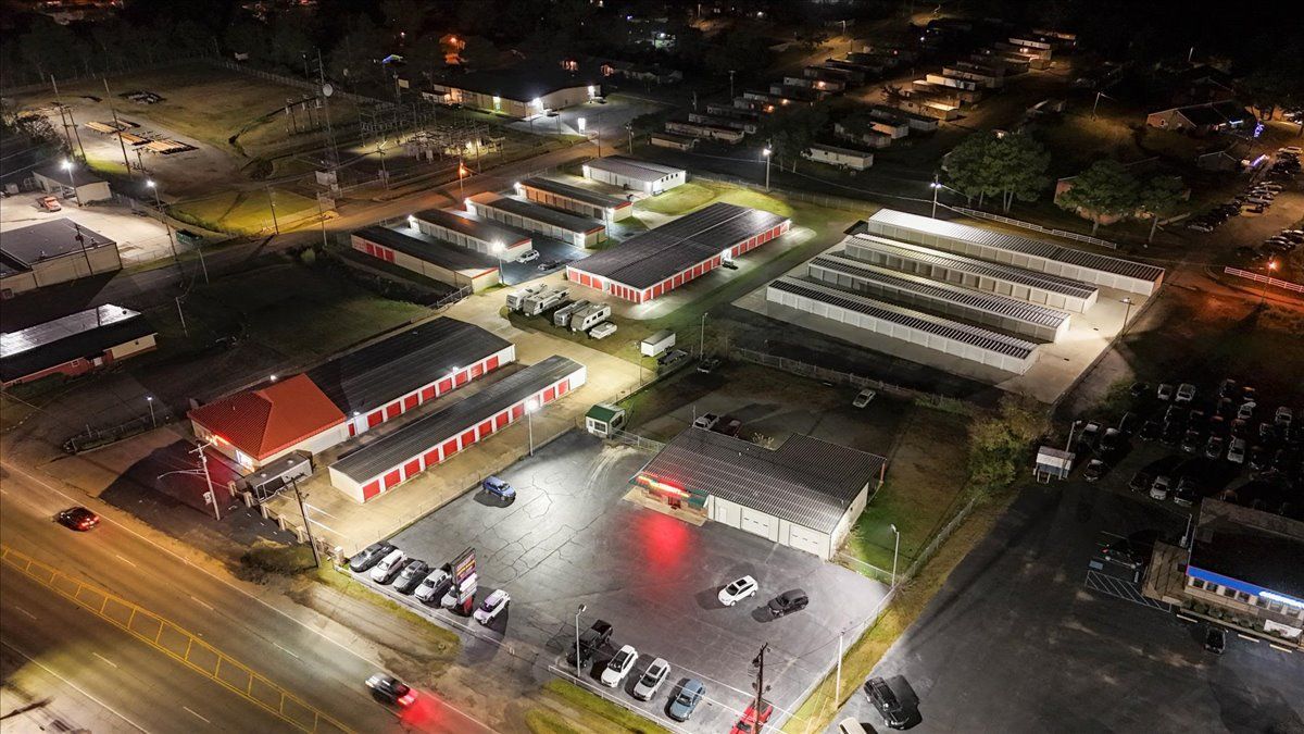 Aerial view of illuminated commercial buildings at night, including storage units and car lot.