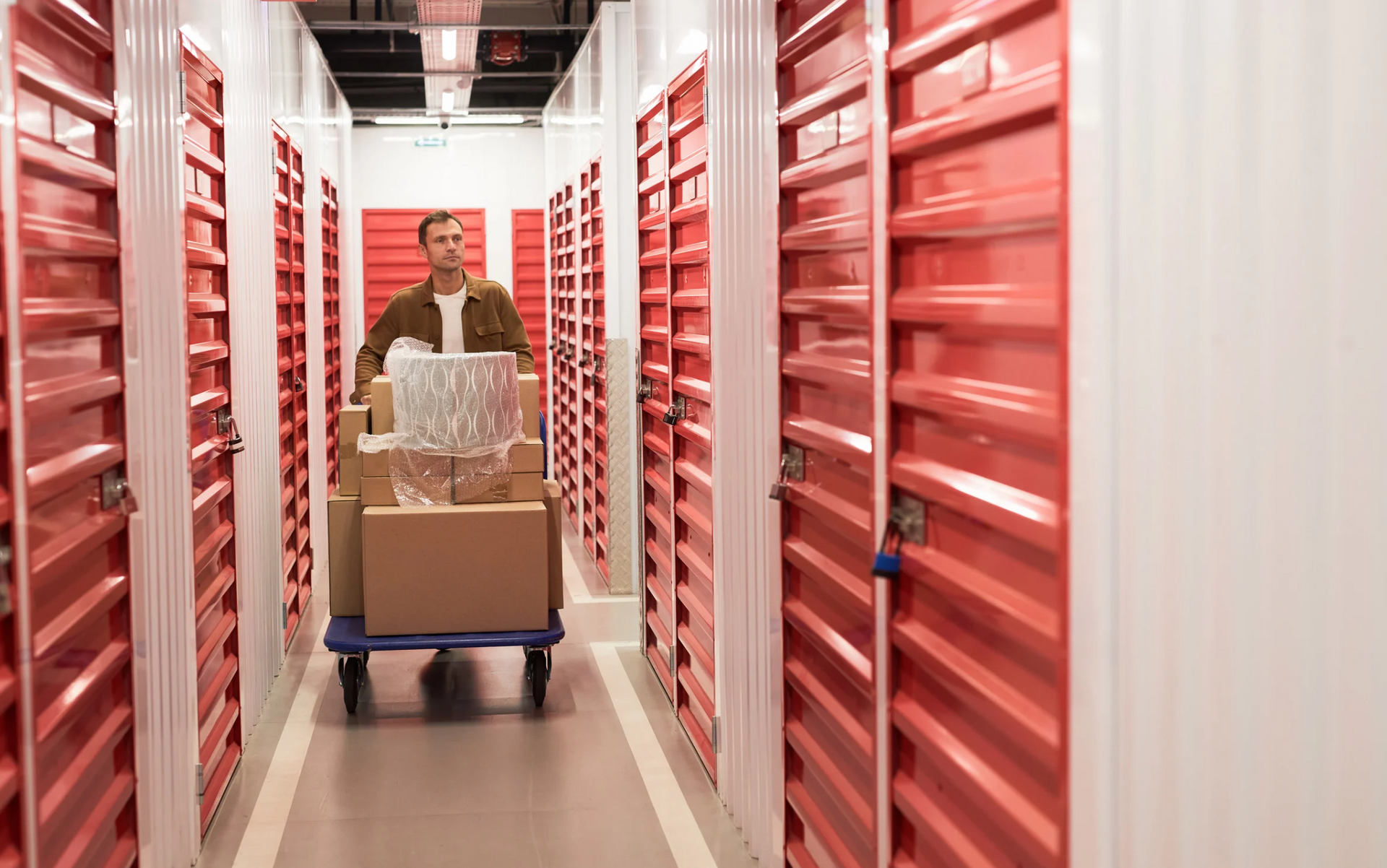 Man pushing a cart of boxes and a lamp down a storage unit hallway with red doors.