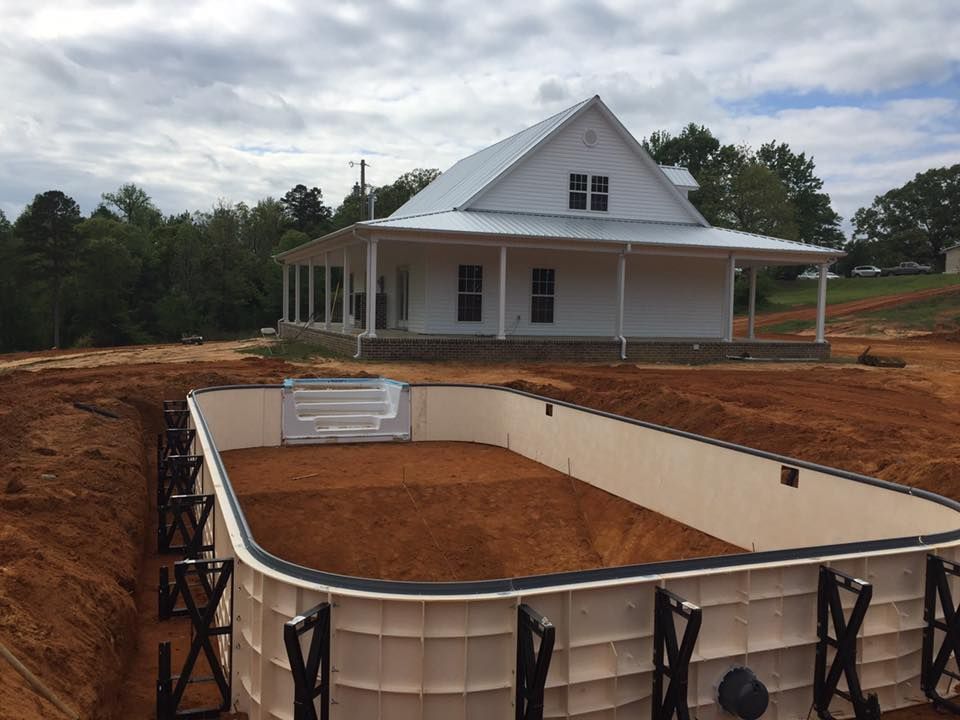 An unfinished rectangular swimming pool frame sits in a dirt backyard in front of a white farmhouse.