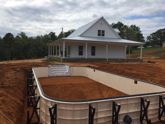 An unfinished rectangular swimming pool frame sits in a dirt backyard in front of a white farmhouse.
