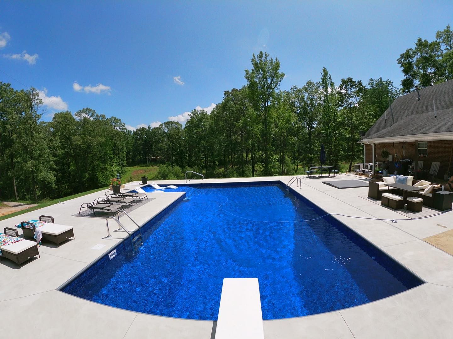 A rectangular swimming pool with deep blue water, a diving board, and patio seating, surrounded by trees under a blue sky.