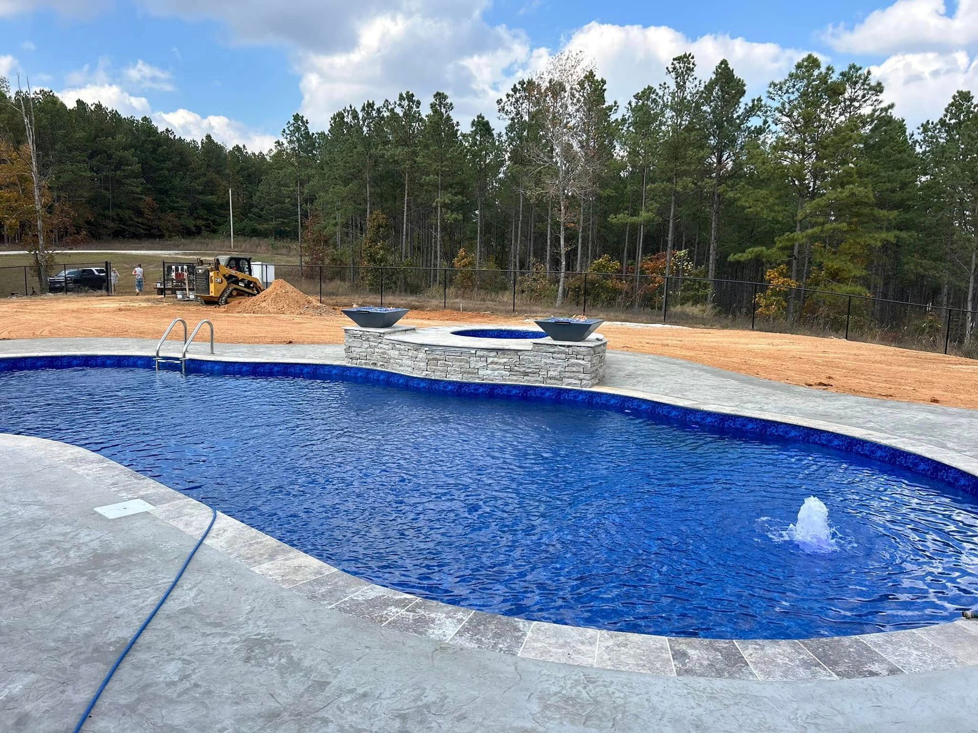 A bright blue swimming pool with a stone spa and fountain sits on a concrete patio in a partially landscaped wooded area.
