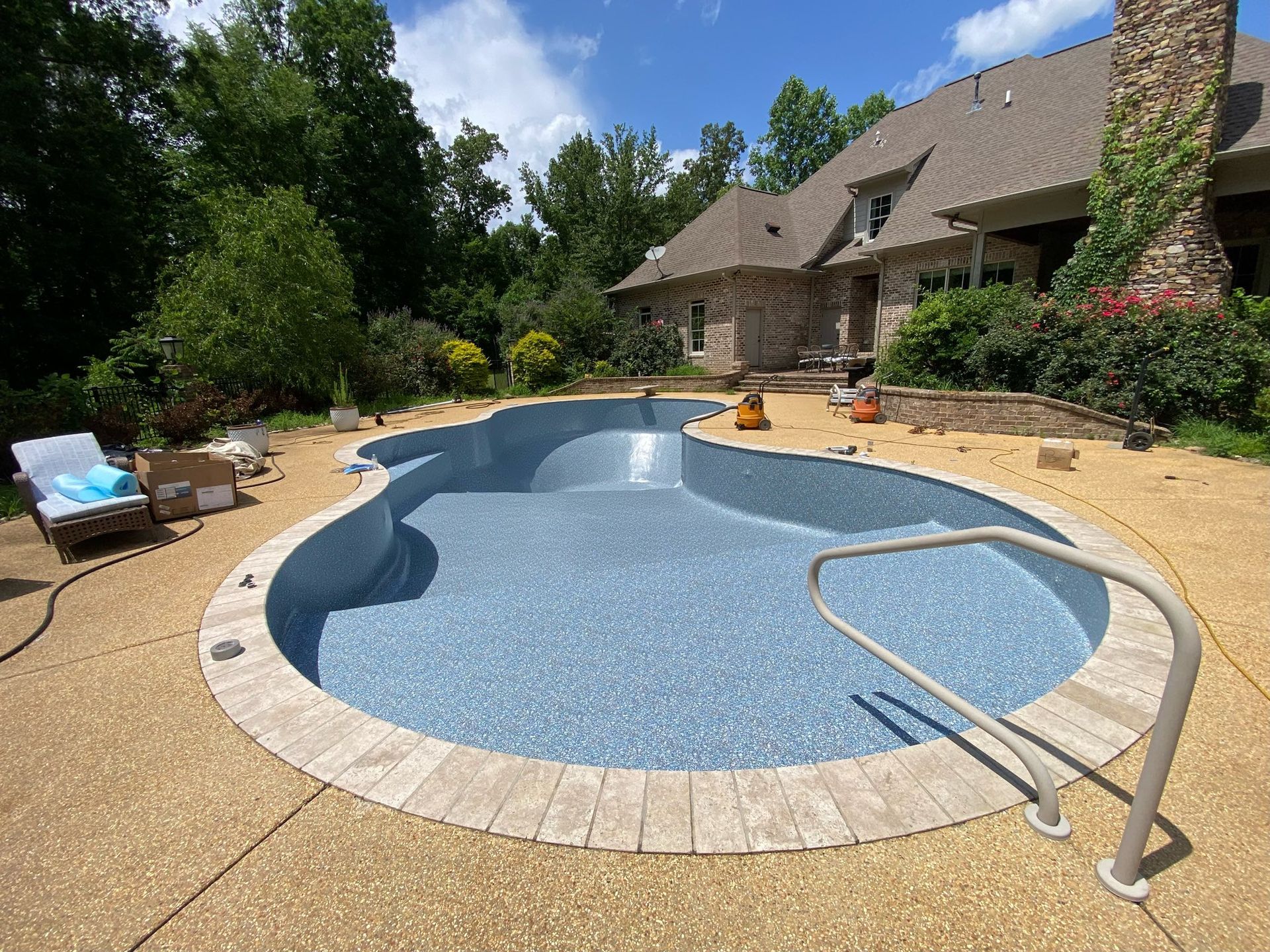 An empty, free-form pool with blue tile finish, surrounded by light-colored textured decking and a stone house.