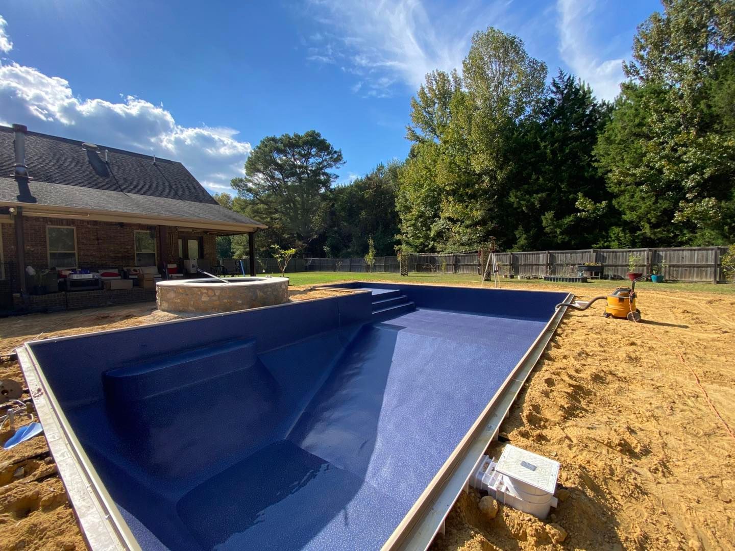 A large, rectangular blue pool shell installed in a dirt backyard next to a house with a stone fire pit.