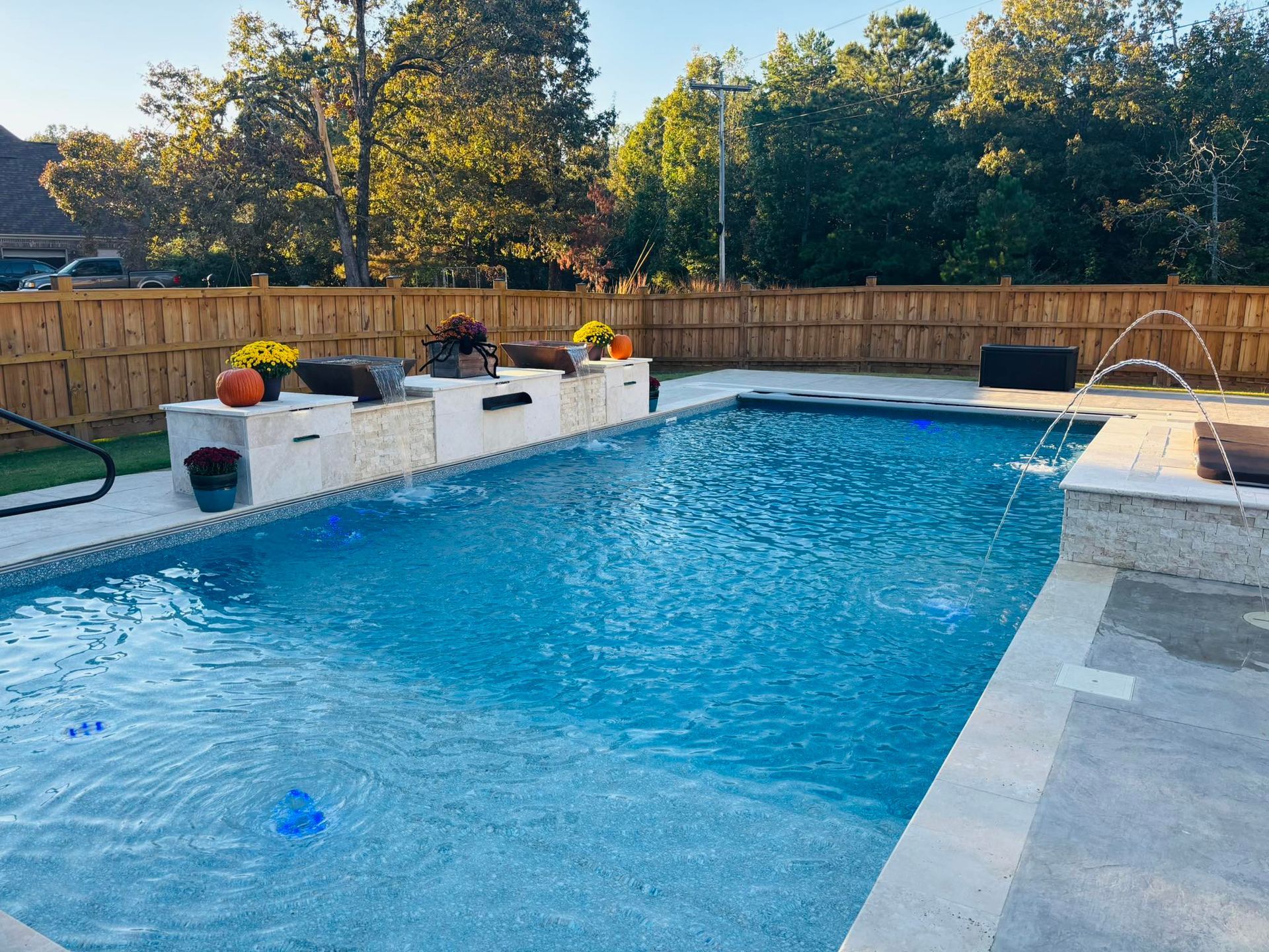 A rectangular backyard swimming pool with fountains, a stone wall with planters, and a wooden fence on a sunny day.