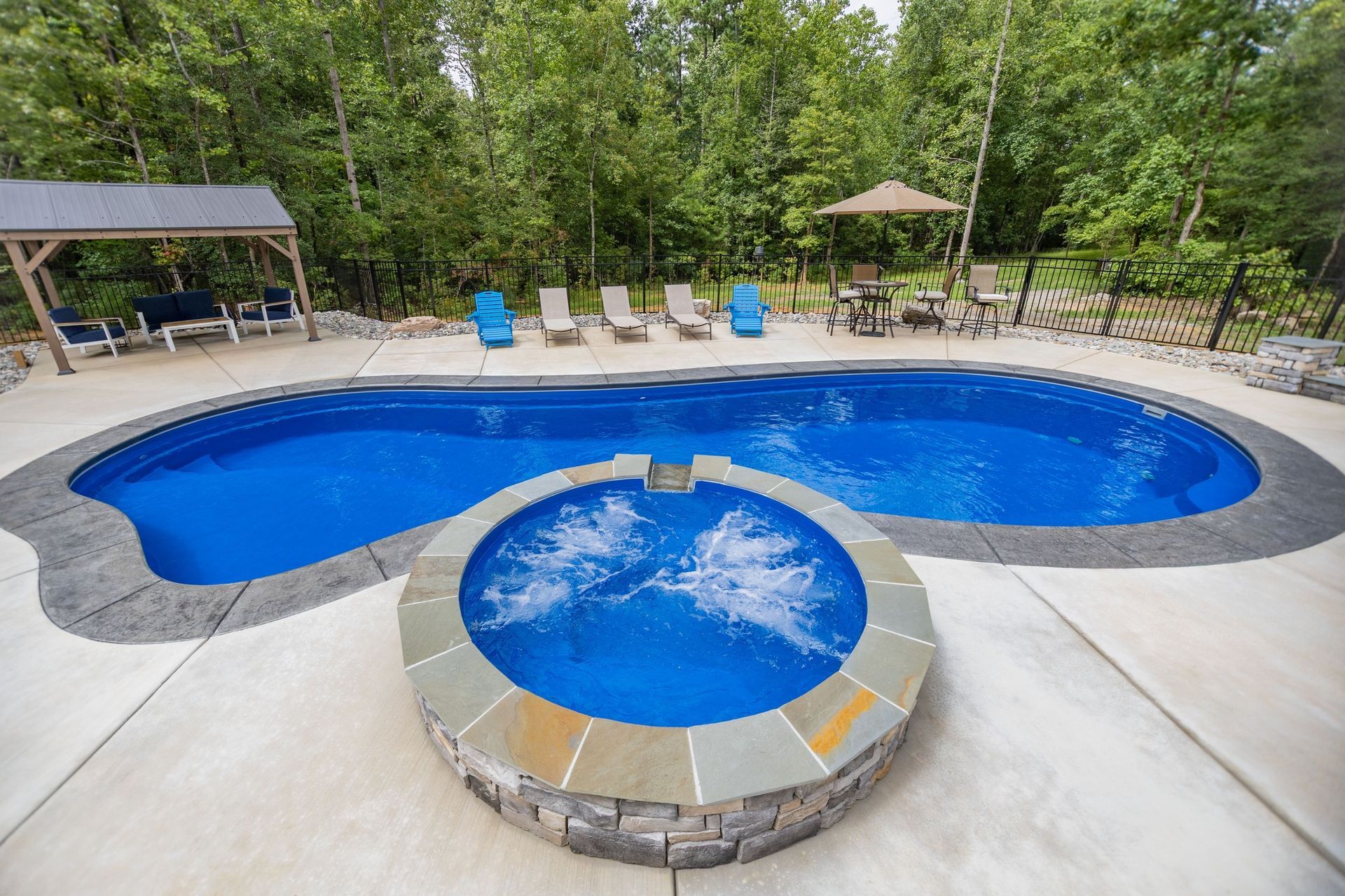 A backyard swimming pool with an attached circular hot tub, surrounded by a patio, loungers, and a wooded forest background.