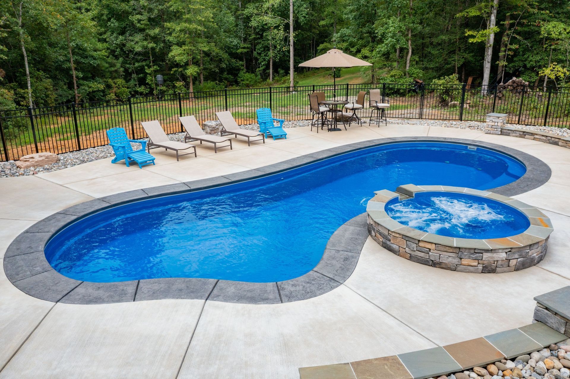 A bright blue kidney-shaped pool with an attached stone hot tub, surrounded by a concrete patio and outdoor chairs.