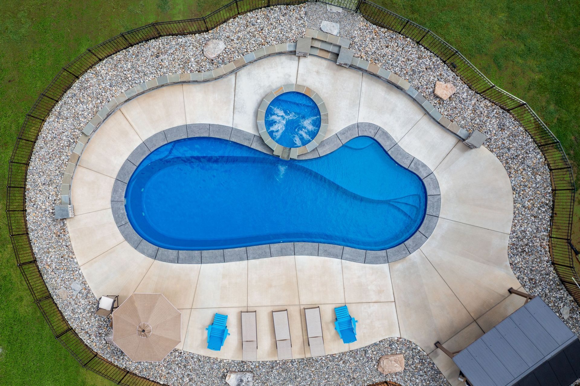 Aerial view of a kidney-shaped swimming pool with an attached circular hot tub, surrounded by a patio and stone border.