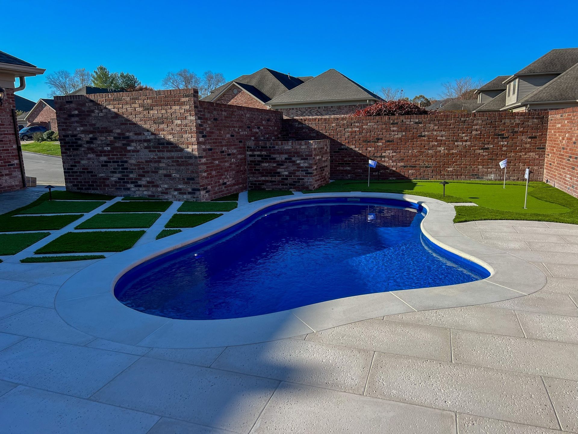 A kidney-shaped blue pool surrounded by a light-colored stone patio, a small putting green, and a red brick wall.