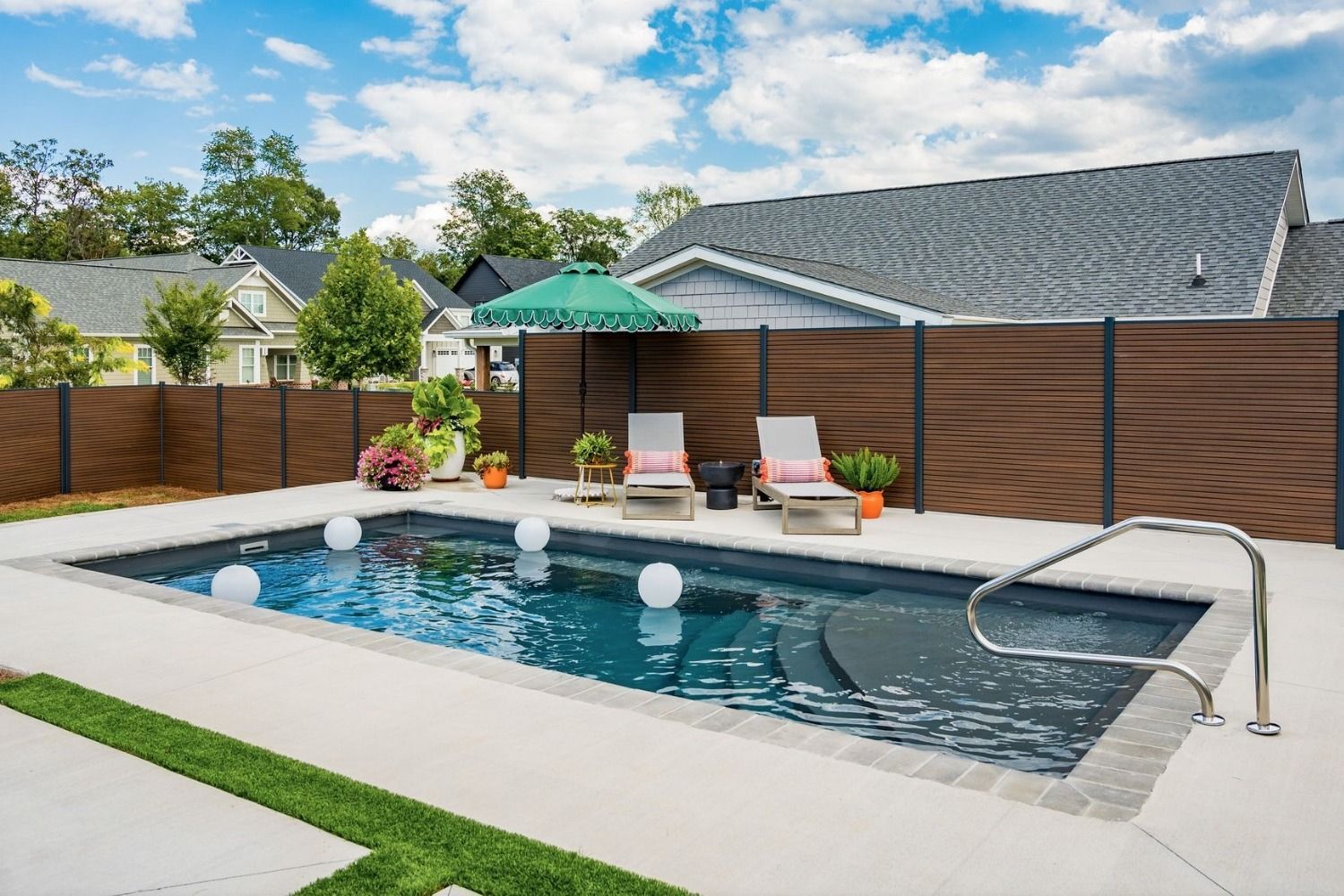 A rectangular inground pool with dark water and built-in steps, surrounded by a patio, a wooden privacy fence, and chairs.