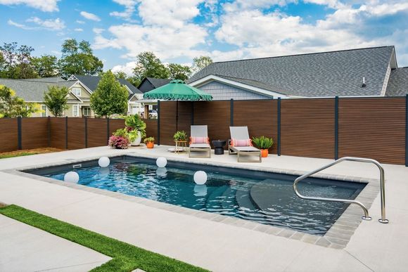 A rectangular inground pool with dark water and built-in steps, surrounded by a patio, a wooden privacy fence, and chairs.