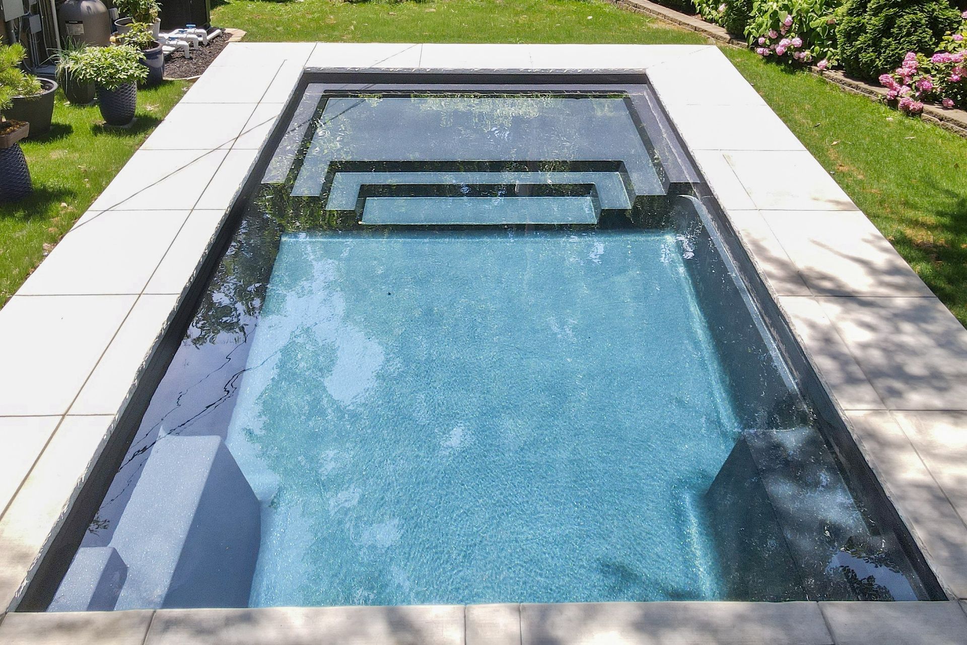 A rectangular, light-blue tiled pool with tiered steps at one end, surrounded by light-colored paving stones and grass.