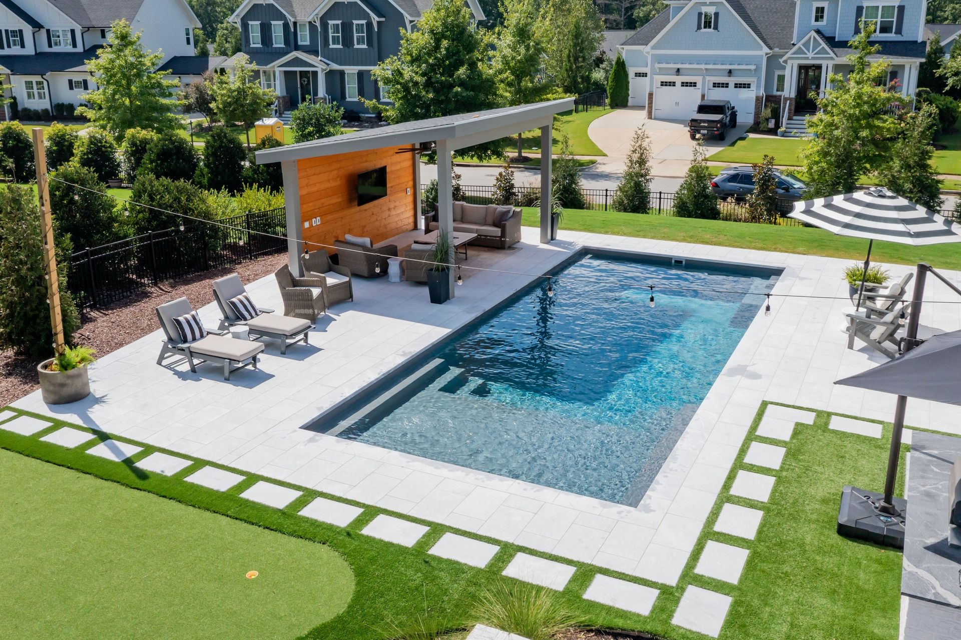 A modern backyard with a rectangular pool, a covered patio with outdoor seating, and a putting green on a sunny day.