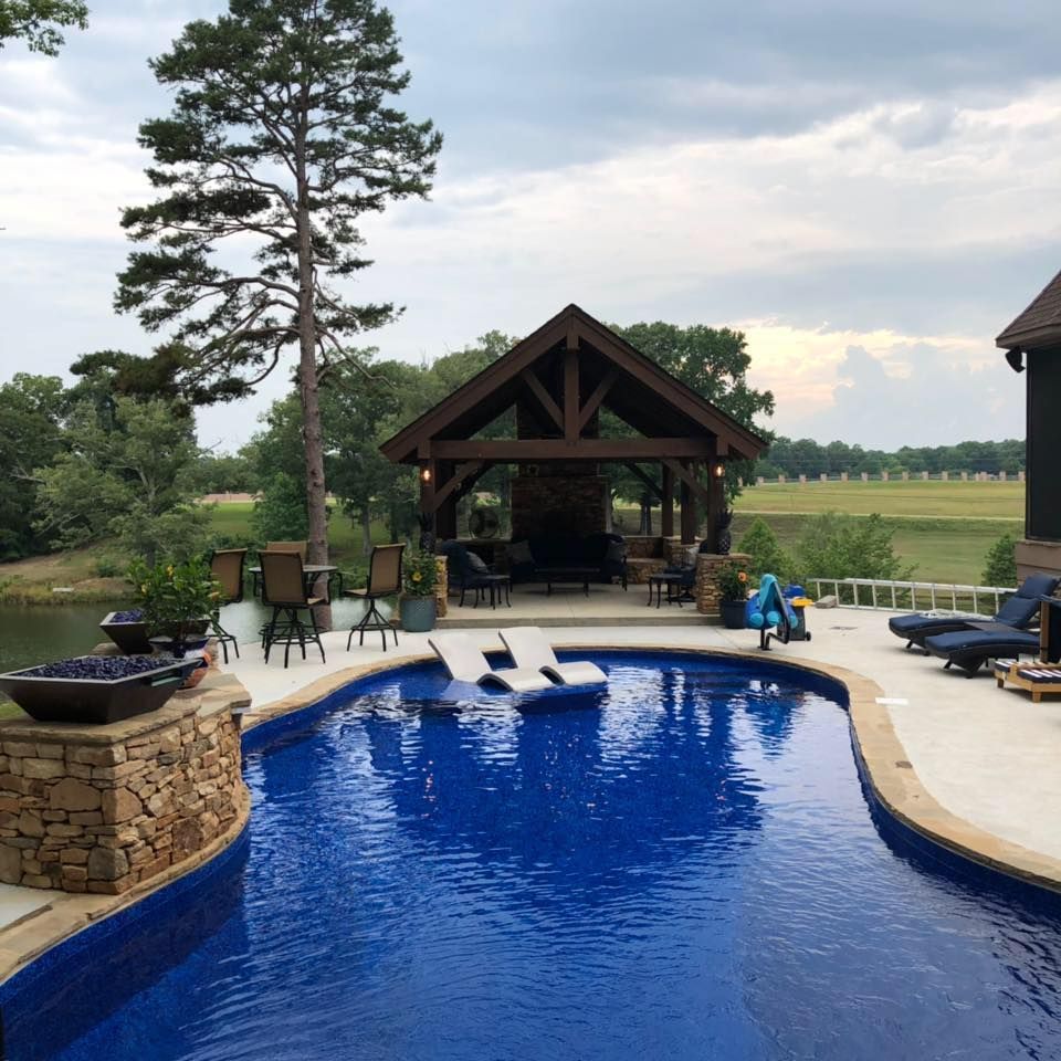 A blue swimming pool with lounge chairs leads to a stone pavilion under a large tree overlooking a grassy field.