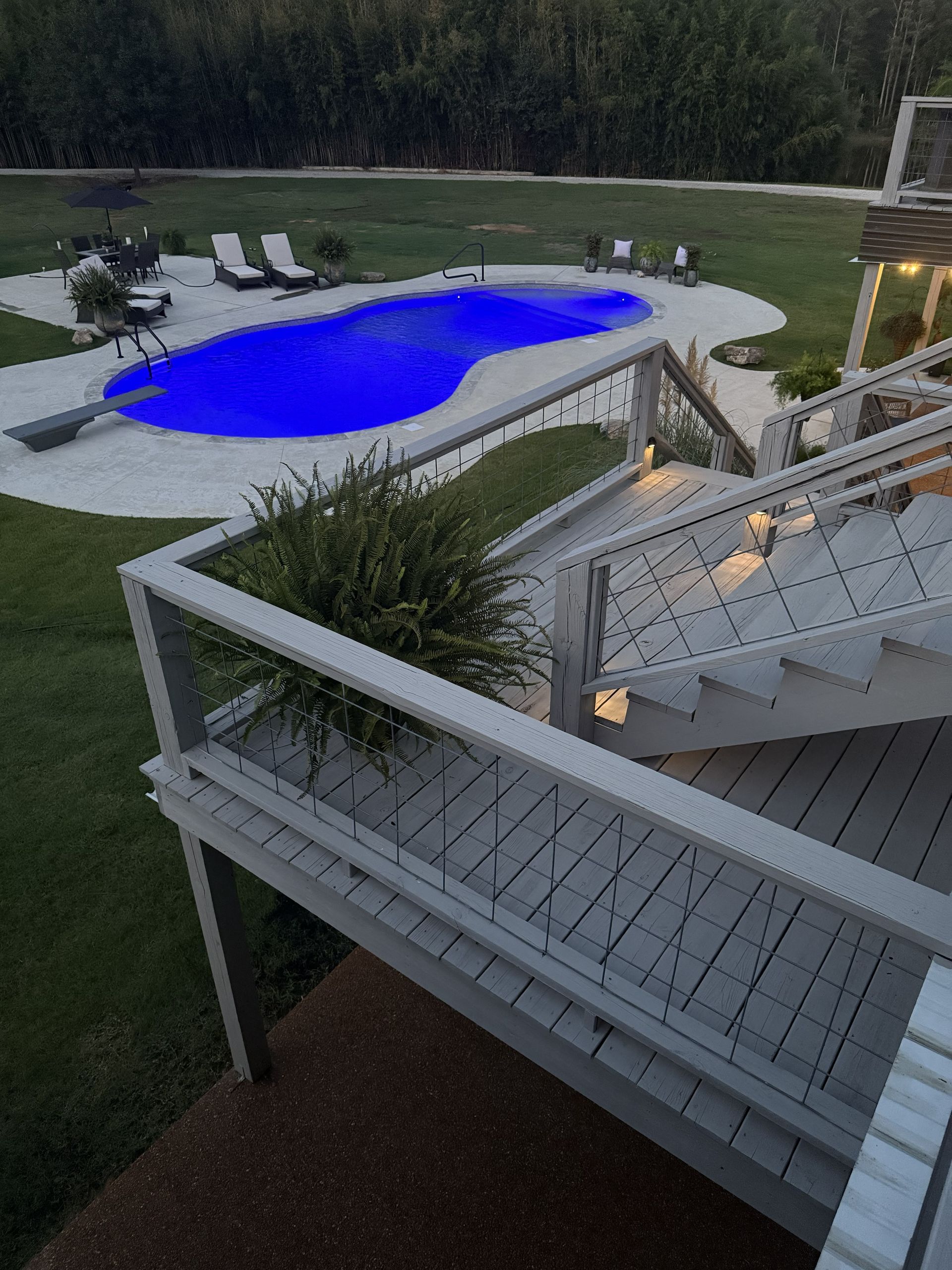 A view from a grey elevated deck looking down at a blue-lit kidney-shaped pool and patio in a grassy backyard at dusk.