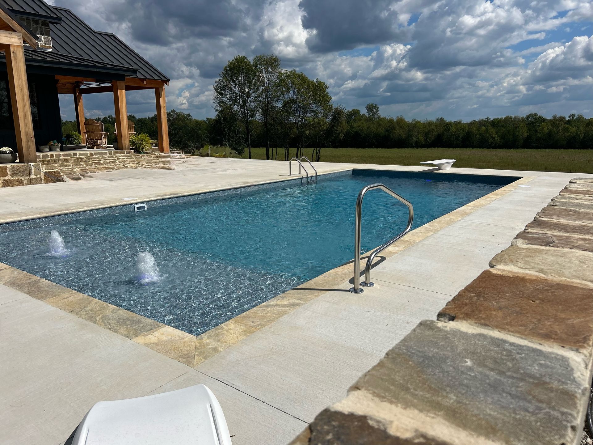 A rectangular swimming pool with fountains and a metal handrail, set on a stone patio beside a home against a cloudy sky.
