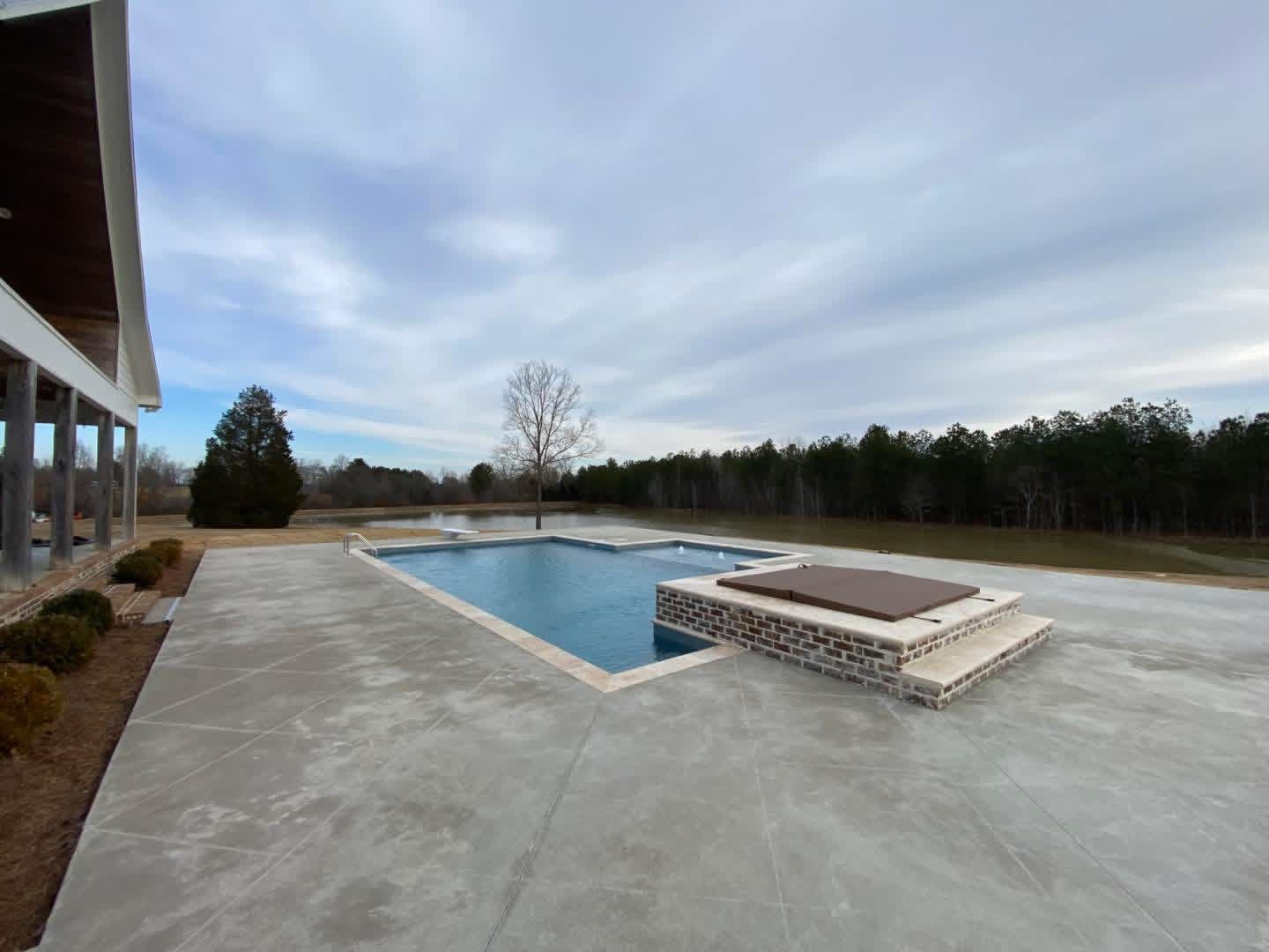 A backyard swimming pool and hot tub on a concrete patio, overlooking a large pond and tree line under a cloudy sky.