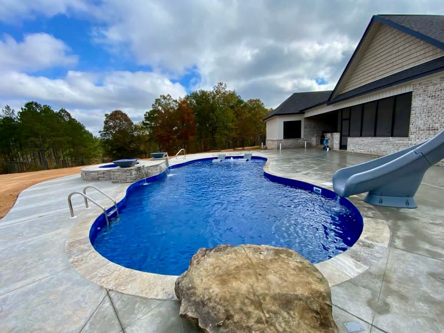 A backyard swimming pool with a stone fountain, a slide, and a hot tub on a concrete patio overlooking trees.