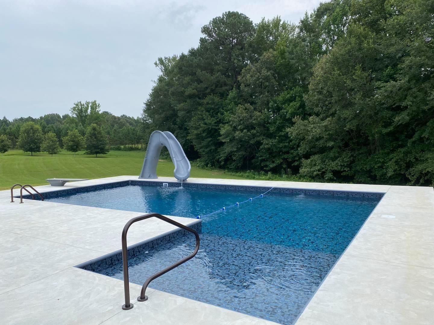 A rectangular inground pool with a slide, a shallow entry step section, and a stone deck, surrounded by trees and grass.