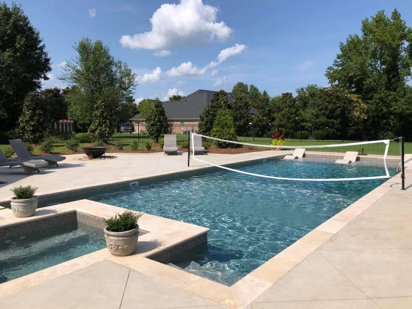 A rectangular swimming pool with a volleyball net, surrounded by a light-colored concrete patio and green landscaping.