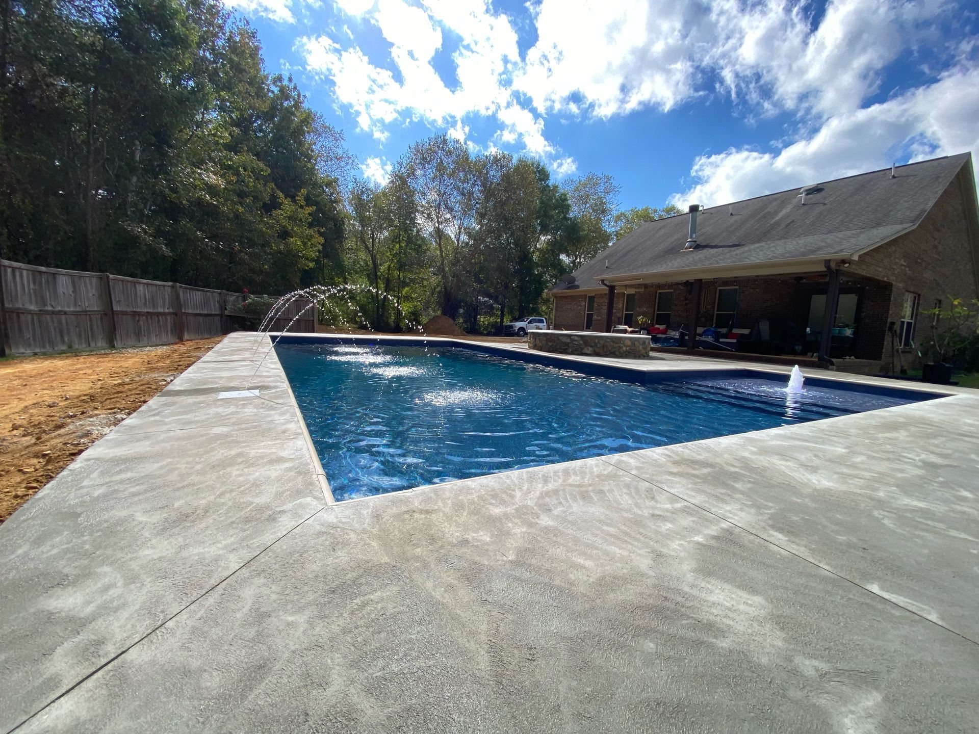 A modern rectangular swimming pool with water jets sits on a concrete patio next to a house under a sunny blue sky.