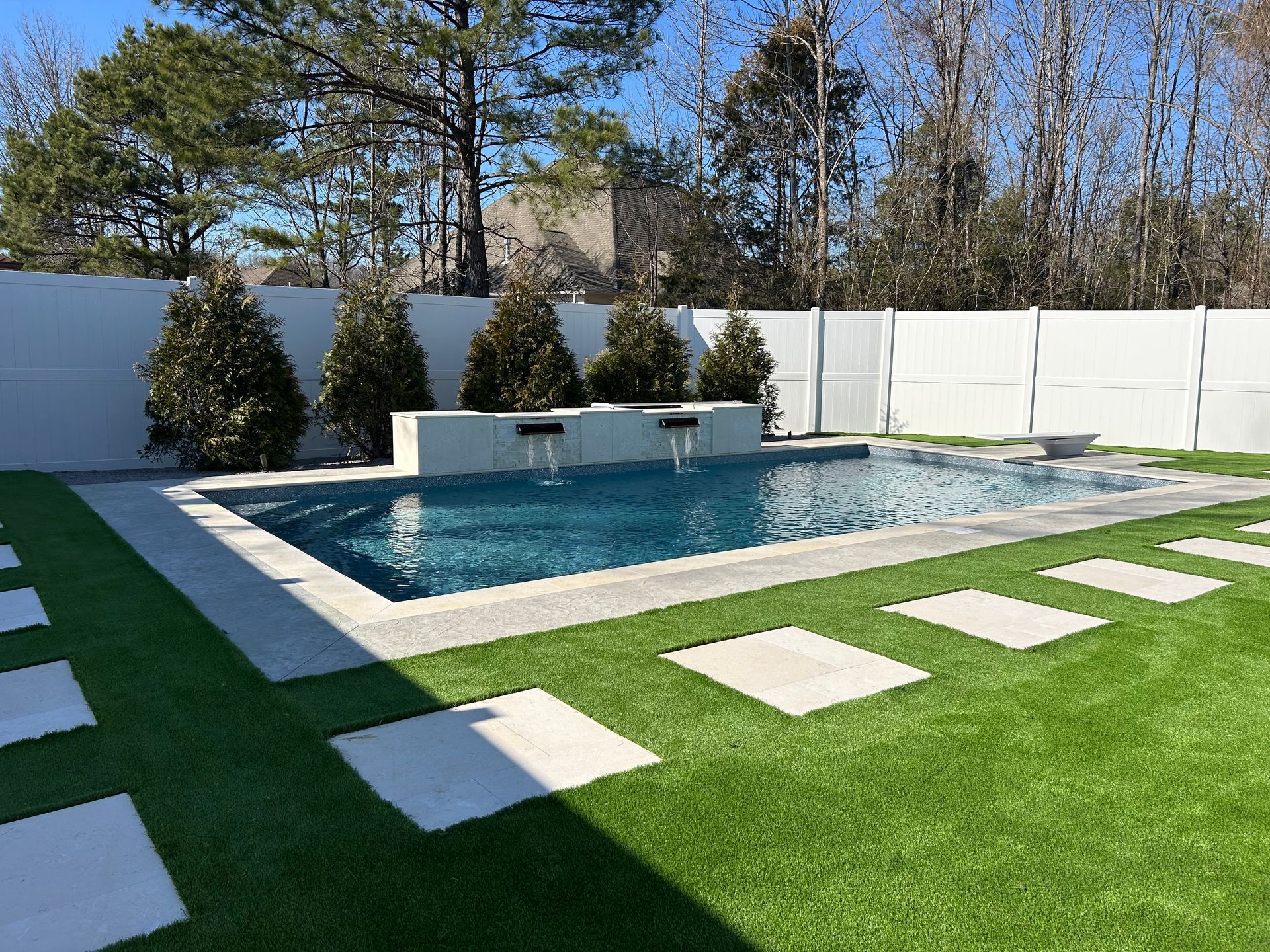 A rectangular swimming pool with water features, surrounded by green turf with stone pavers and a white privacy fence.