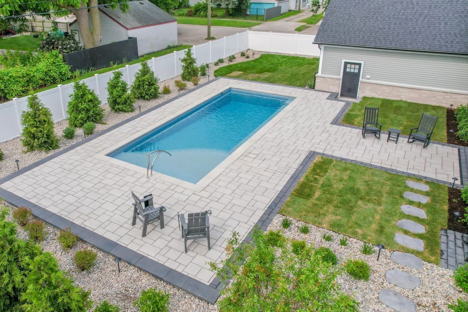 An outdoor rectangular swimming pool surrounded by a light-colored paver patio, a small lawn, and landscaping.