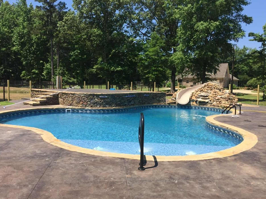 A kidney-shaped swimming pool with a stone wall feature, slide, and patio in a backyard setting with trees.