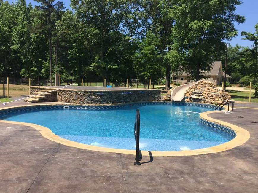 A kidney-shaped swimming pool with a stone wall feature, slide, and patio in a backyard setting with trees.