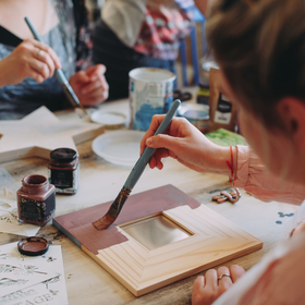 People paint wooden frames and decorative shapes with brushes and jars of brown paint on a table.
