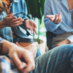 Three people sit in a group discussion, gesturing while talking, with one holding a takeaway coffee cup.