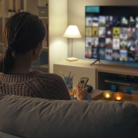A person sitting on a sofa in a dimly lit room, holding a remote control while looking at a large television screen.