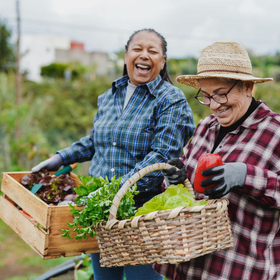 Two people in plaid shirts and gardening gloves hold baskets of fresh produce and smile while standing in a garden.