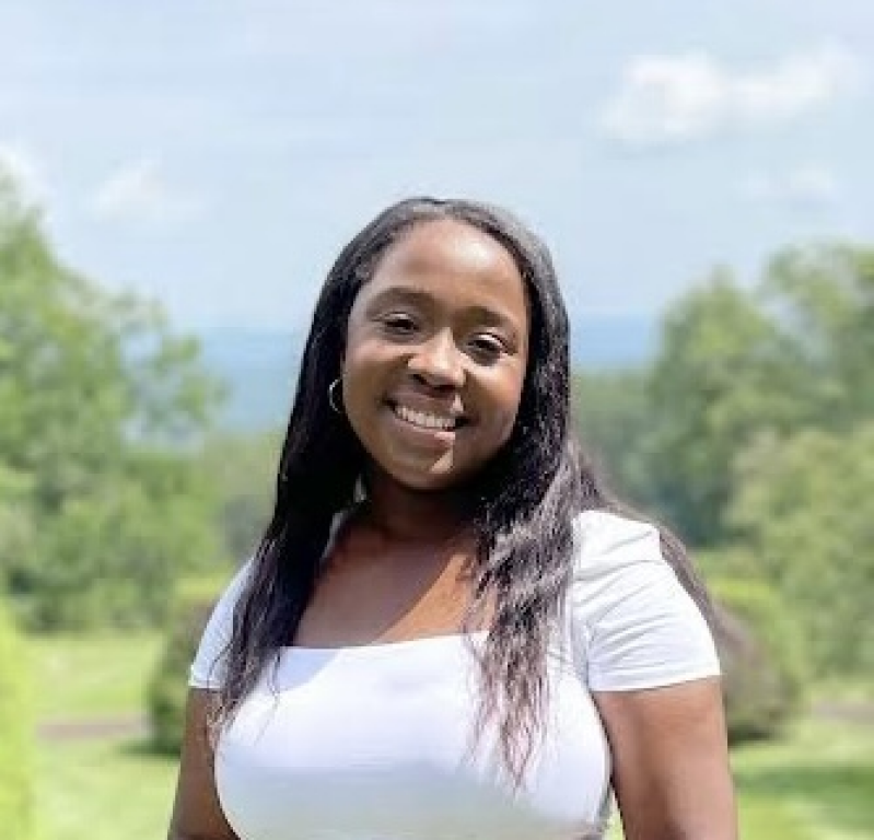 Smiling person in a white top standing outdoors with green trees and mountains in the background