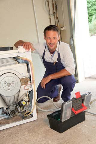 Repairman kneeling beside a washing machine, smiling. He is wearing a blue overall and holding a toolbox.