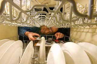 Man inside a dishwasher reaching for items on a rack, surrounded by plates.