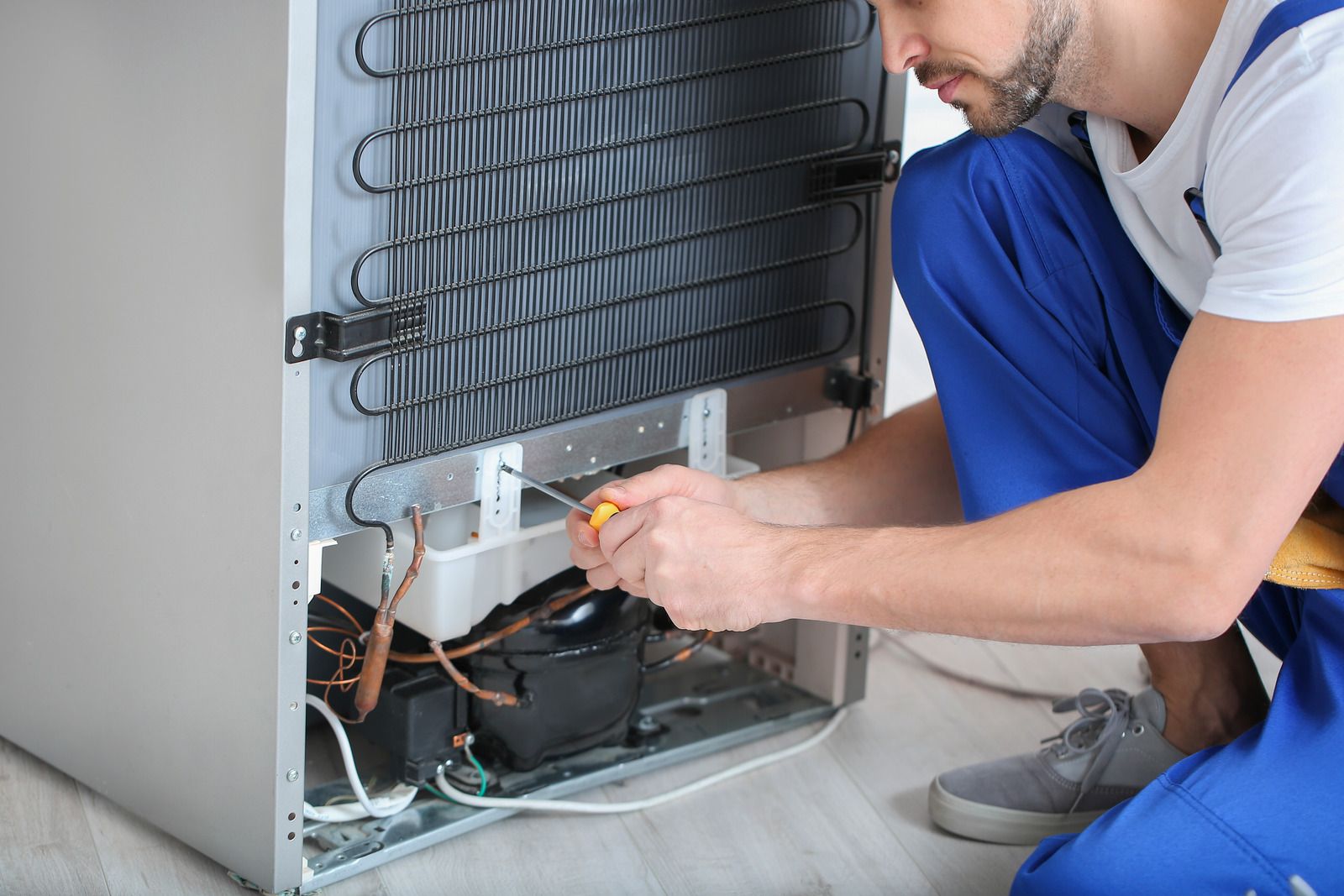 Man in blue overalls repairs a refrigerator's back, using a screwdriver; interior wires and compressor visible.
