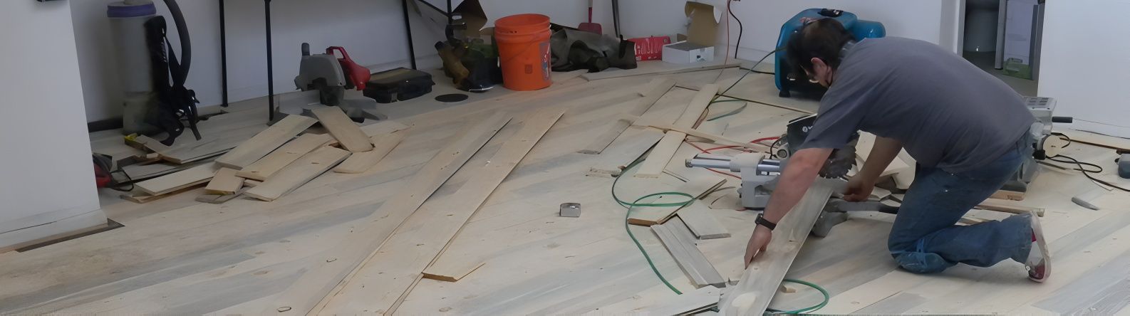 Person installing wooden planks on floor. Construction site with tools, a bucket, and white walls.