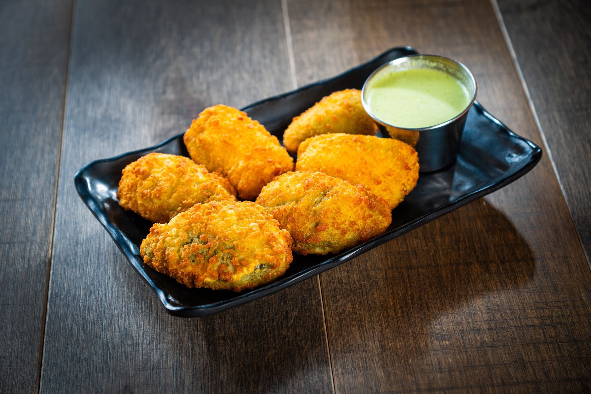 Golden fried snacks on a black platter with green dipping sauce on a wooden table