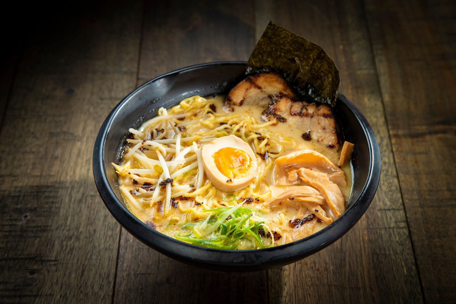 Bowl of ramen with egg, sliced pork, noodles, and green onions on a dark wooden table