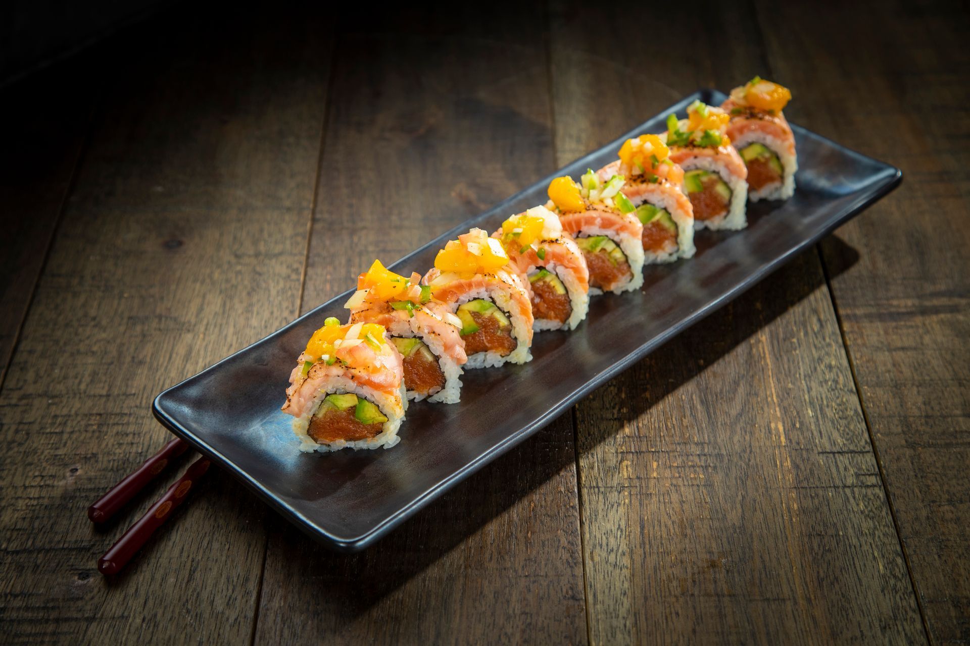 Sushi rolls on a black rectangular plate with chopsticks, served on a dark wooden table.