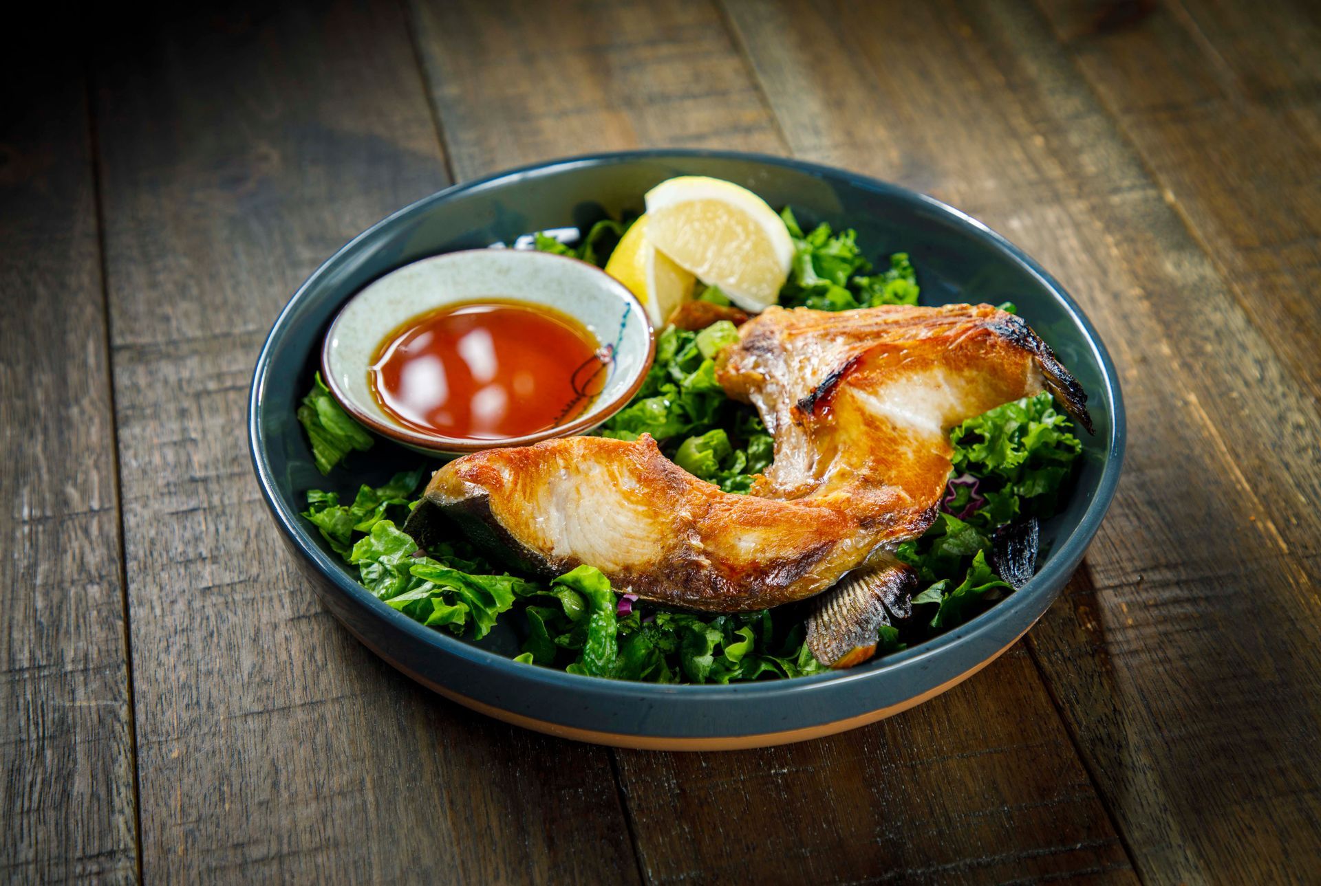 Roasted chicken leg on a bowl of greens with lemon wedges and dipping sauce on a wooden table