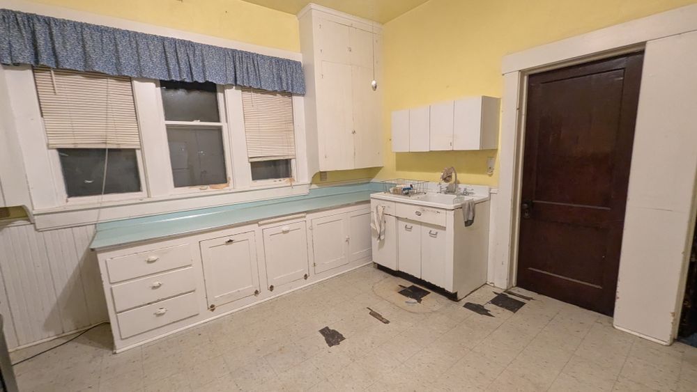 Old, yellow kitchen with white cabinets, a blue countertop, and a dark brown door.