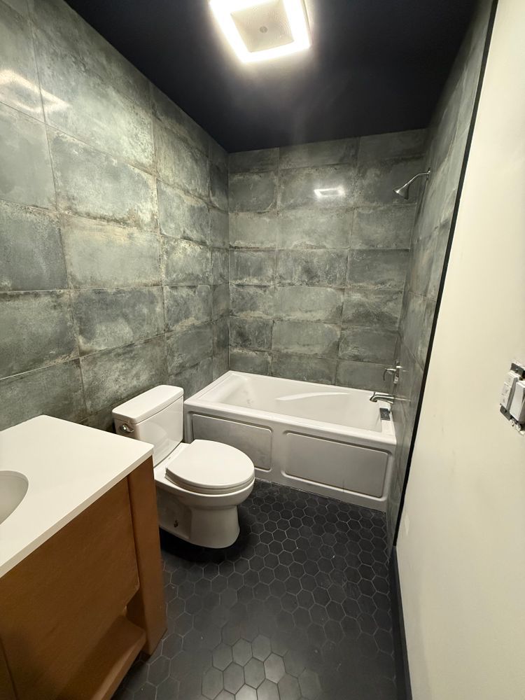 Modern bathroom with gray-tiled walls, black hexagonal floor, white tub, toilet, and vanity with light wood.