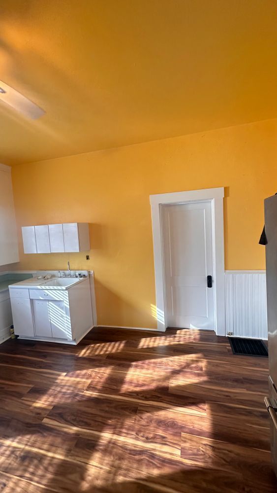 Yellow-walled kitchen with white cabinets, door, and wood-look flooring. Sunlight streams in.