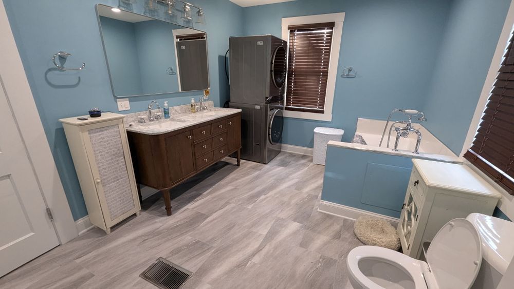 A bathroom with a light blue wall, dark wooden vanity, white toilet, and a washing machine next to the window.