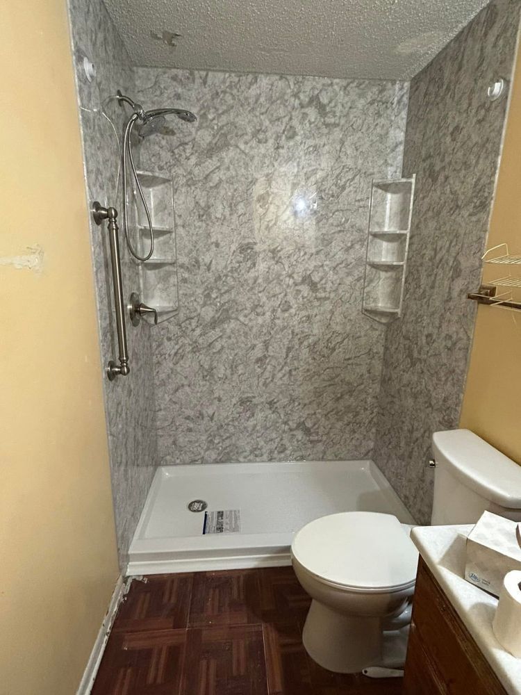 Shower with marble-look walls, white base, and shelves. Toilet and sink in a bathroom with wood-look flooring.