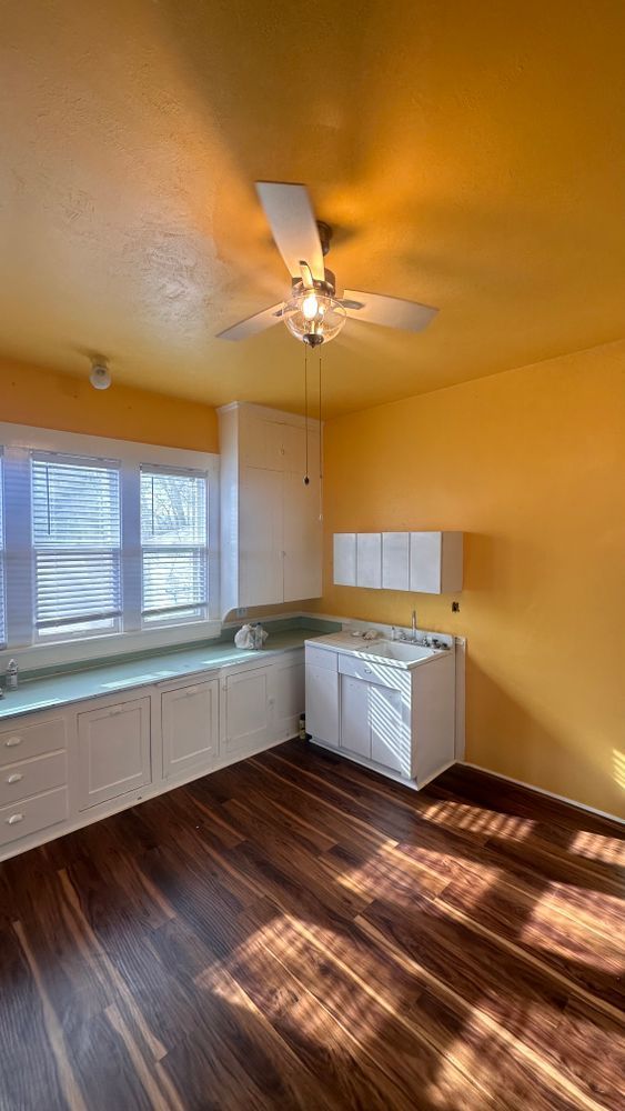 A small kitchen with yellow walls, white cabinets, and dark wood floors. A window and a ceiling fan are visible.