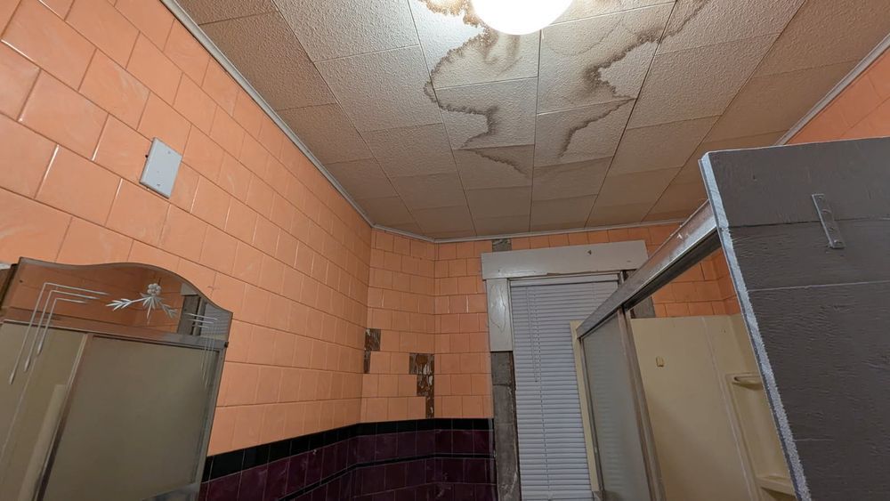Orange-tiled public restroom with water-damaged ceiling. Stalls and a sink are visible.