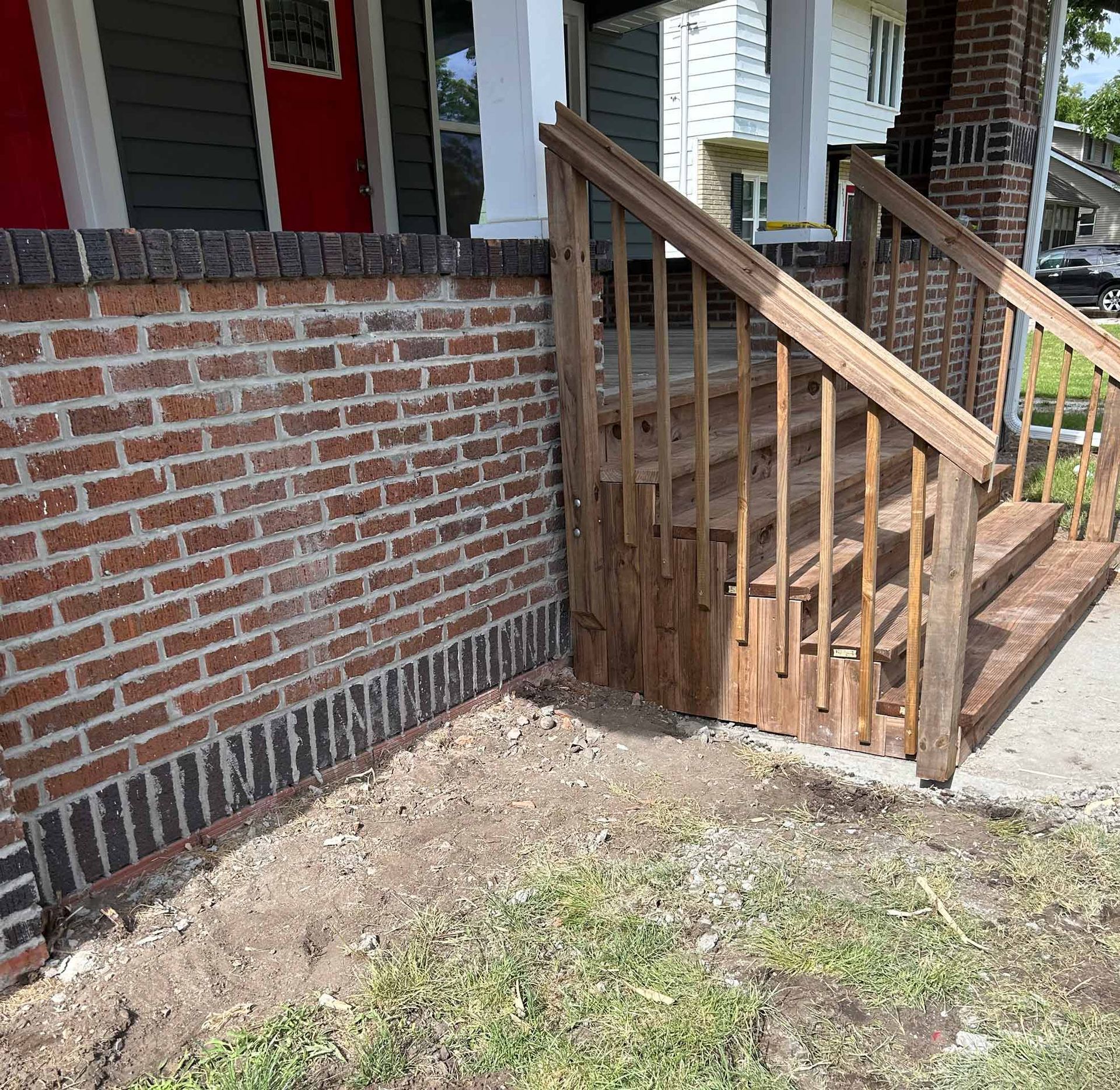 Brick wall and wooden steps leading to a house porch. Soil and grass in the foreground.